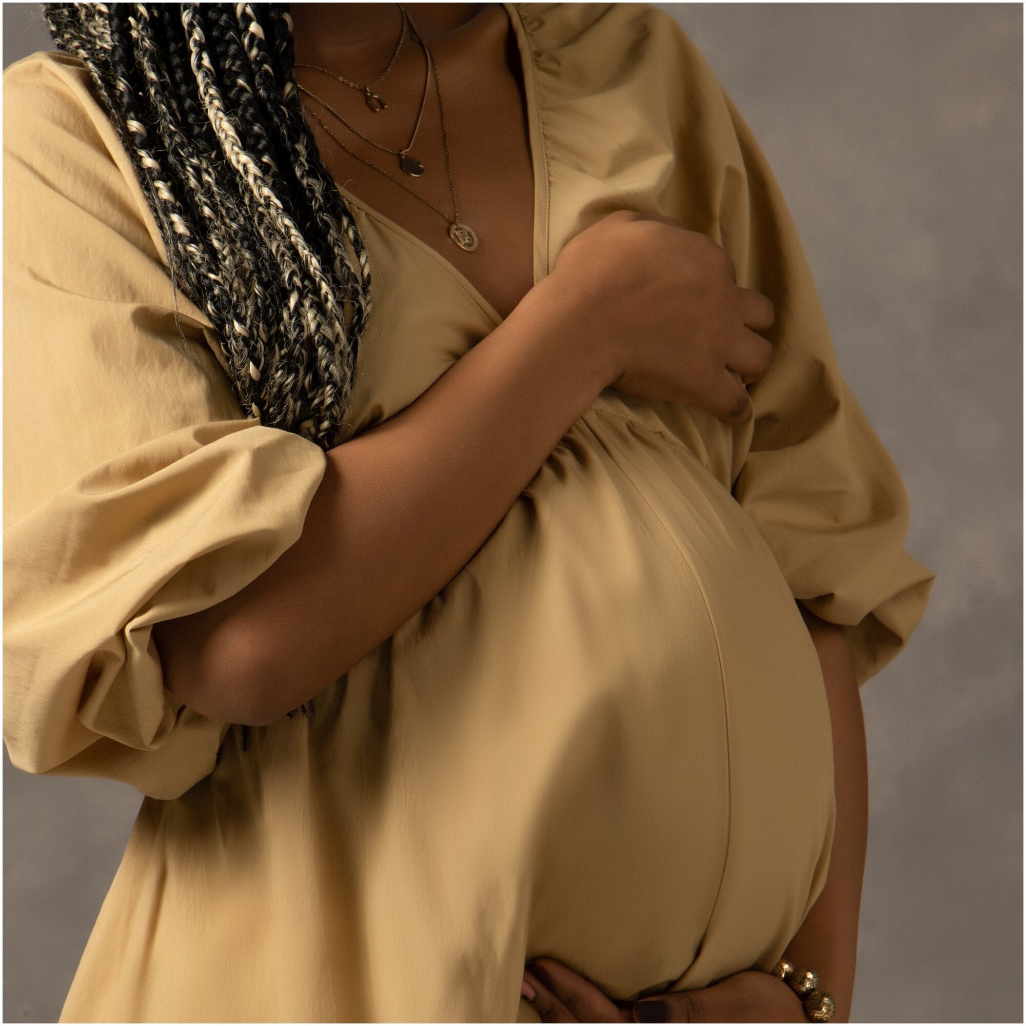 Rasta haired pregnant woman is holding her stomach wearing a cushy, beige dress.